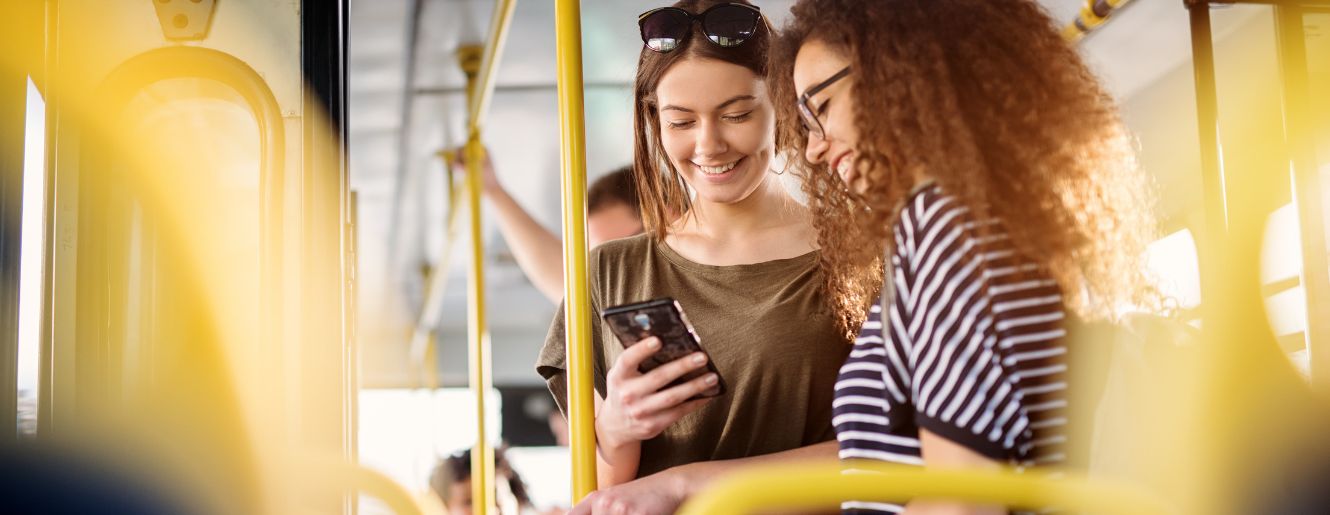 two female presenting people looking at a phone while standing on a bus